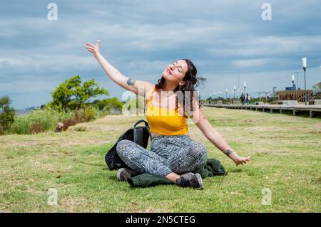 beautiful latin young woman sitting sunbathing with her eyes closed and content sitting cross-legged on the grass smiling, dressed in yellow, arms out Stock Photo