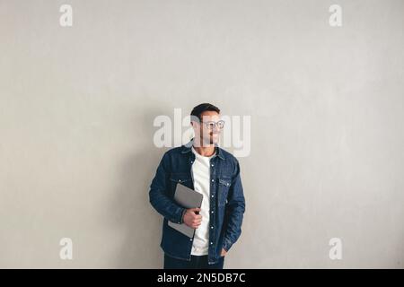 Young businessman looking away while standing against a grey wall with a laptop in his hand. Happy businessman wearing business casual and eyeglasses Stock Photo