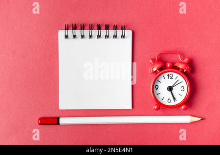 Top view of open empty spiral notebook and alarm clock with pen on red background. Stock Photo