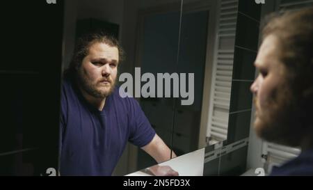 Dramatic person standing in front of bathroom mirror looking at reflection suffering from problems. One pensive serious overweight man facing himself Stock Photo