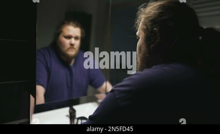 Dramatic person standing in front of bathroom mirror looking at reflection suffering from problems. One pensive serious overweight man facing himself Stock Photo