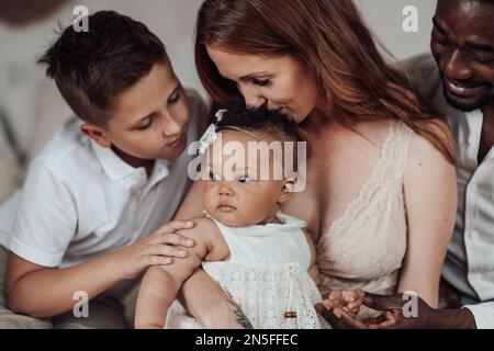 Mixed race family with white son and infant swarthy daughter spend time together at cozy apartment. African american man with his fair skinned wife ki Stock Photo