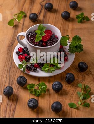 A small white cup on a saucer and wooden cutting board is filled with berries and green leaves Stock Photo