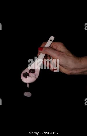 A female hand holding a ceramic spoon with berries inside and dripping yoghurt, on a black background Stock Photo