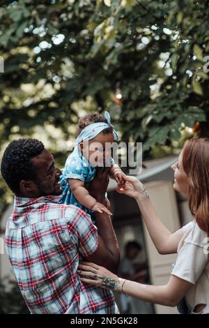 Mixed race family hold dark skinned infant daughter in air spend time together near travel trainer. African american man his fair skin wife and little Stock Photo