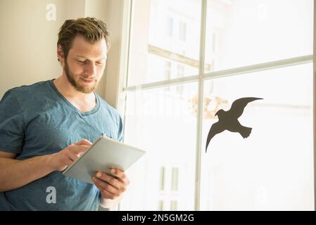 Man using digital tablet standing by window, Munich, Bavaria, Germany Stock Photo