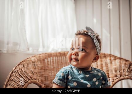 Swarthy little girl in blue polka dot dress has positive emotion sitting on cane chair light and shade on face. African american baby infant relaxes a Stock Photo