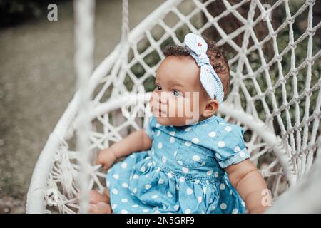 Swarthy little girl in blue polka dot dress has positive emotion sitting on white macrame cotton rope cocoon hanging. African american baby infant rel Stock Photo