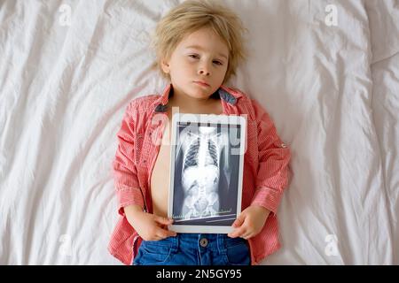 Toddler child, blond boy, holding x-ray picture on tablet of child body with swallowed magnet showing, child swallow dangerous object Stock Photo