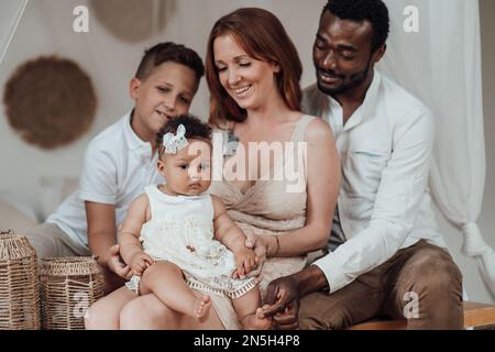 Mixed race family with white son and infant swarthy daughter spend time together at cozy apartment. African american man with his fair skinned wife ki Stock Photo