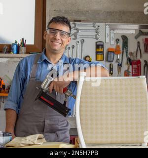 Smiling upholsterer in his workshop with various tools and nail gun after restoring an old chair. Stock Photo