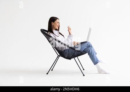 Cheery Caucasian woman in headphones having online video call on laptop, sitting on chair against white wall Stock Photo
