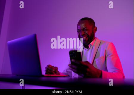 the man is happy at the computer. a dark-skinned man works in computer. neon light. A man works at a computer at night Stock Photo