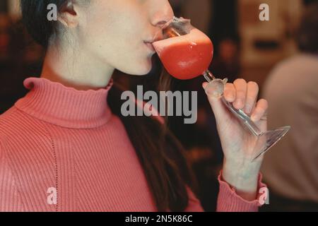 A close-up photo of a female hand holding a pink cocktail glass in the shape of a rose. The hand belongs to a woman wearing a pink turtleneck sweater, Stock Photo