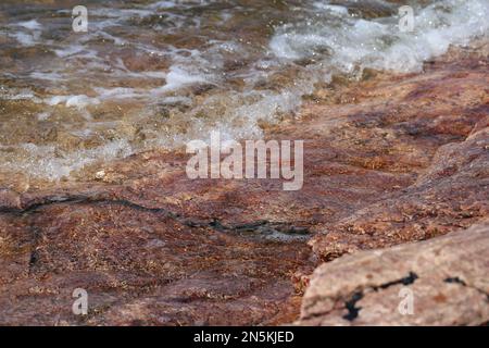 Lots of small waves hitting red and black beach rocks in Eira aka Eiranranta, Helsinki, Finland. Sunny summer day by the Baltic Sea. Color image. Stock Photo