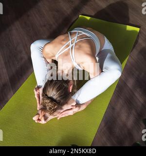 Young woman practicing yoga, doing kurmasana exercise, a variation of the turtle pose, exercising alone, sitting on a mat in white sportswear in the s Stock Photo