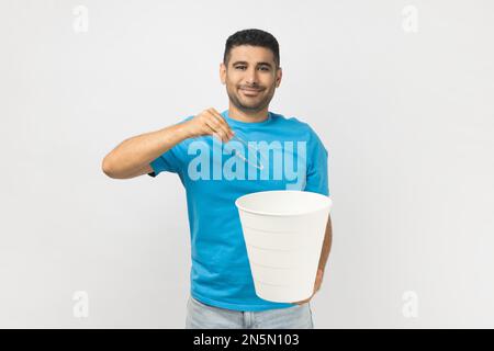 Portrait of satisfied delighted unshaven man wearing blue T- shirt standing after laser eyesight correction, throwing away his eyeglasses. Indoor studio shot isolated on gray background. Stock Photo