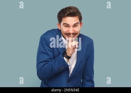 Portrait of funny smiling handsome man with mustache standing with finger near lips showing shh gesture, keeps secret wearing official style suit. Indoor studio shot isolated on light blue background. Stock Photo