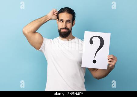 Portrait of puzzled man with beard wearing white T-shirt holding question mark, finding smart solution, asking for advice, looking away. Indoor studio shot isolated on blue background. Stock Photo