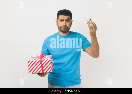 Portrait of puzzled attractive man wearing blue T- shirt standing choosing between two present boxes, choosing little gift box with disappointment. Indoor studio shot isolated on gray background. Stock Photo