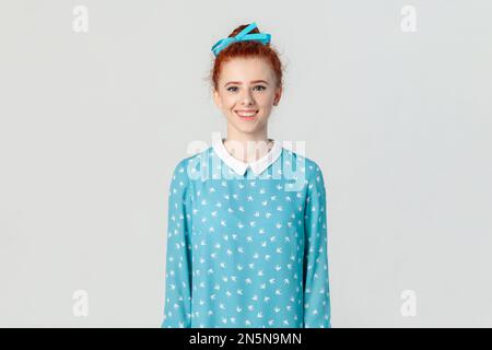 Portrait of smiling red haired woman with bun hairstyle, standing looking at camera with happy positive expression, wearing blue dress. Indoor studio shot isolated on gray background. Stock Photo