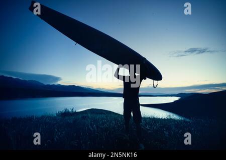 Man in silhouette holding SUP board on his head near lake at sunset in Kazakhstan. Stand up paddle boarding outdoor active recreation in nature. Stock Photo
