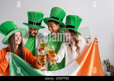cheerful and interracial friends in green hats holding glasses of beer and Irish flag while celebrating Saint Patrick Day Stock Photo