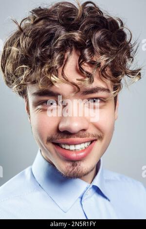 Portrait of attractive man with beard and mustache with wavy hairstyle, wearing blue shirt, looking at camera with toothy smile. Indoor studio shot isolated on gray background. Stock Photo