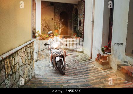 Cheerfully smiling man in helmet and sunglasses riding the motor scooter on the narrow Sicilian old town streets. Happy Italian vacation and transport Stock Photo