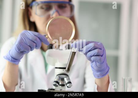 Scientist stands near microscope and looks at test tube with solution through magnifying glass. Stock Photo