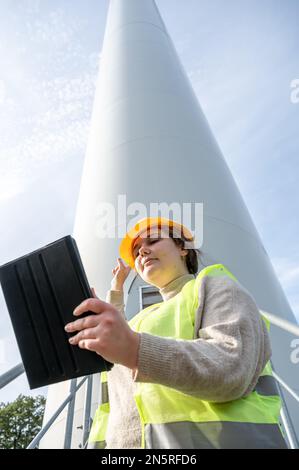 Female engineer with brown curly hair and yellow helmet using her digital tablet while standing in front of a wind turbine during daylight, low angle Stock Photo