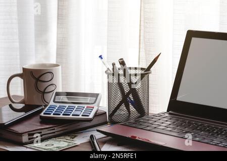 A desktop in front os a window where are a laptop, calculator, agenda, documents, pens, money and a mug. Stock Photo