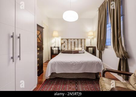 Regal style bedroom with white wardrobe, red rug, dresser with dark drawers, white shell bed and window with hanging curtains Stock Photo