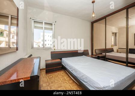 Double bedroom with vintage root furniture combined with black, mattress without bedding and built-in wardrobe with sliding mirror doors Stock Photo