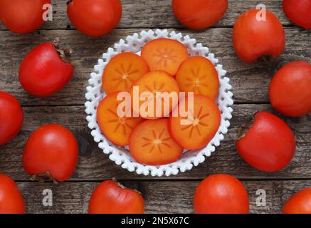 Slices of persimmon fruit in bowl on wooden background, table top view. Fruit background with sliced and whole kaki fruits, directly above photo. Stock Photo
