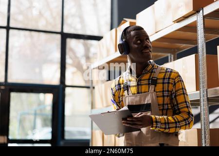 African american storehouse employee in headphones supervising warehouse merchandise. Young industrial warehouse worker listening to music, looking at shelf and taking notes Stock Photo