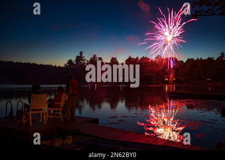 A silhouette of three people sit on a pier at a lake and watch fireworks explode in the night sky on the Fourth of July holiday. Stock Photo