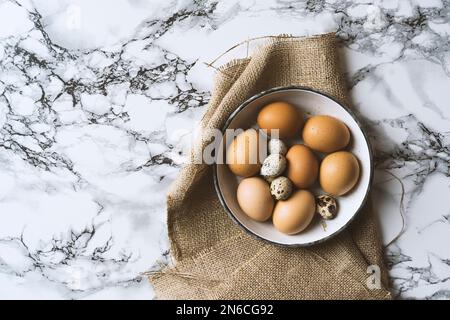 Fresh organic free range chicken eggs and quail eggs in enamel bowl on marble countertop. Top view shot. Stock Photo
