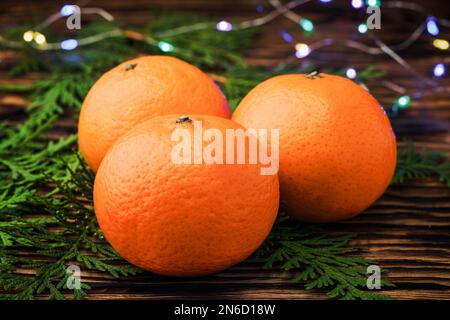 Tangerines close - up on a wooden table near the coniferous branches of juniper on the background of burning lights garlands. Christmas background. Ch Stock Photo
