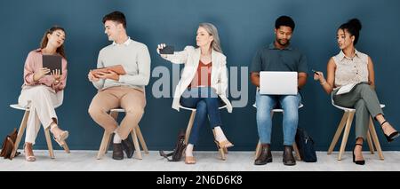 Group of diverse businesspeople sitting on chairs in a row in an office together. Five business professionals sitting and talking using technology in Stock Photo