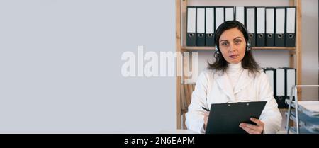 Female doctor wear headset talking to patient and writing notes on clipboard having online video call, webcam view. Telemedicine concept. Banner Stock Photo