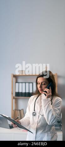 Confident young female doctor calling on her mobile phone while holding a clipboard and standing in the consultation. Copy space. Banner Stock Photo