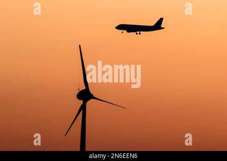 Airplane silhouetted flying past a wind turbine at early dawning. Stock Photo