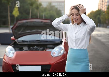 Stressed woman standing near broken car on city street Stock Photo
