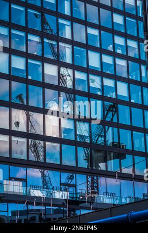 Glass facade of a modern office building reflecting the blue sky and two construction cranes, vertical Stock Photo