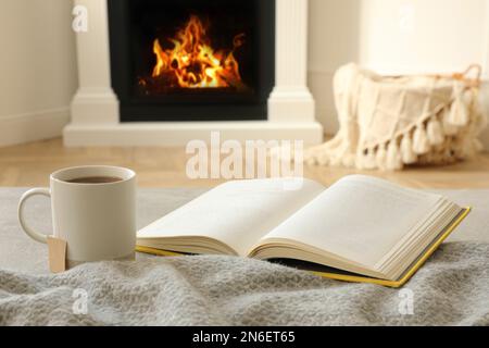 Cup of hot tea and book near fireplace at home. Cozy atmosphere Stock Photo