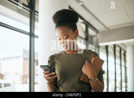 African american business woman texting on smartphone holding journal and walking in modern office. Smiling female entrepreneur using mobile app or Stock Photo