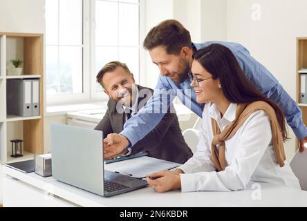 Friendly cheerful colleagues in office together look at laptop screen and discuss business project. Stock Photo
