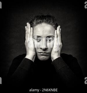 Conceptual dark portrait of unhappy woman with hands over her head stretching face in ugly grimace.  Black and White film grain. Stock Photo