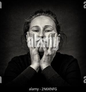 Conceptual dark portrait of old tired woman stretching the skin of her face in ugly grimace.  Black and White film grain. Stock Photo